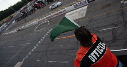 Woman wearing a red shirt, waving a checkered flag over the racing surface.